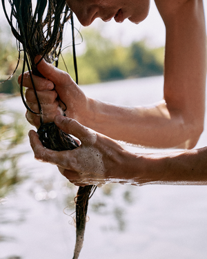 Person washing hair with water outdoors near a body of water
