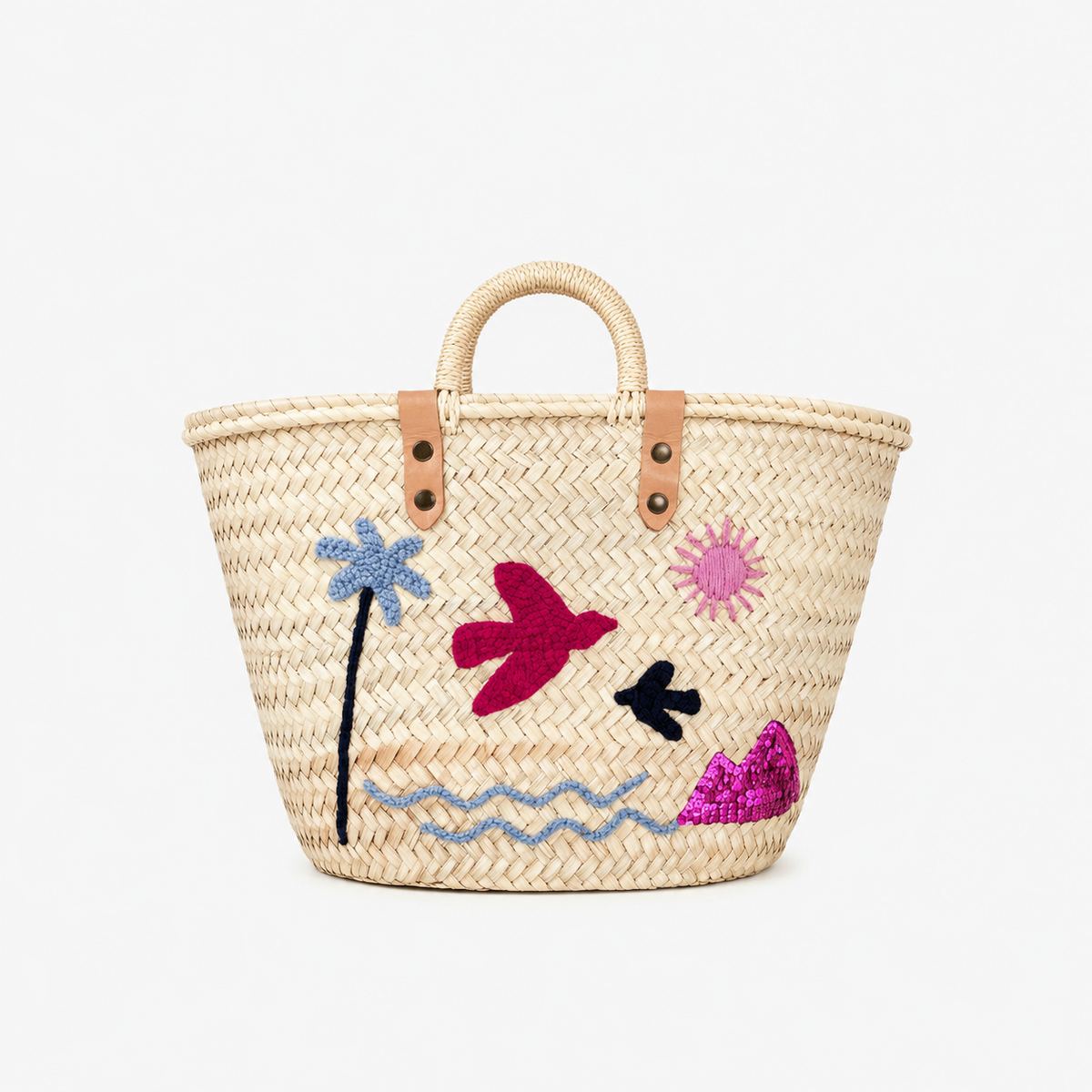 A natural woven straw tote bag with tan leather handles and embroidered tropical motifs including a blue palm tree, pink sun, magenta bird, black bird, and wavy blue line art on a white background.