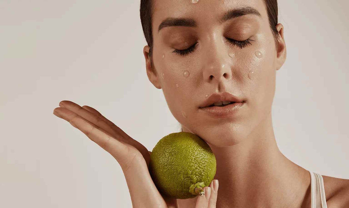 Facial close-up of woman with water droplets on skin holding a green lime near her face with eyes closed