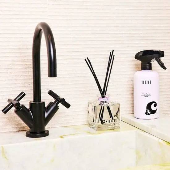 Janzen Room Spray &C Lavender Rose & Relax on a bathroom sink next to a black faucet and diffuser sticks.