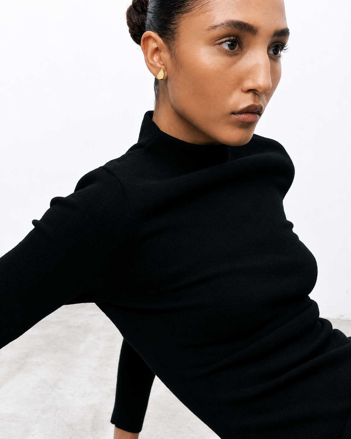 A close-up of a model wearing a black mock neck long-sleeve knit top against a light gray background.
