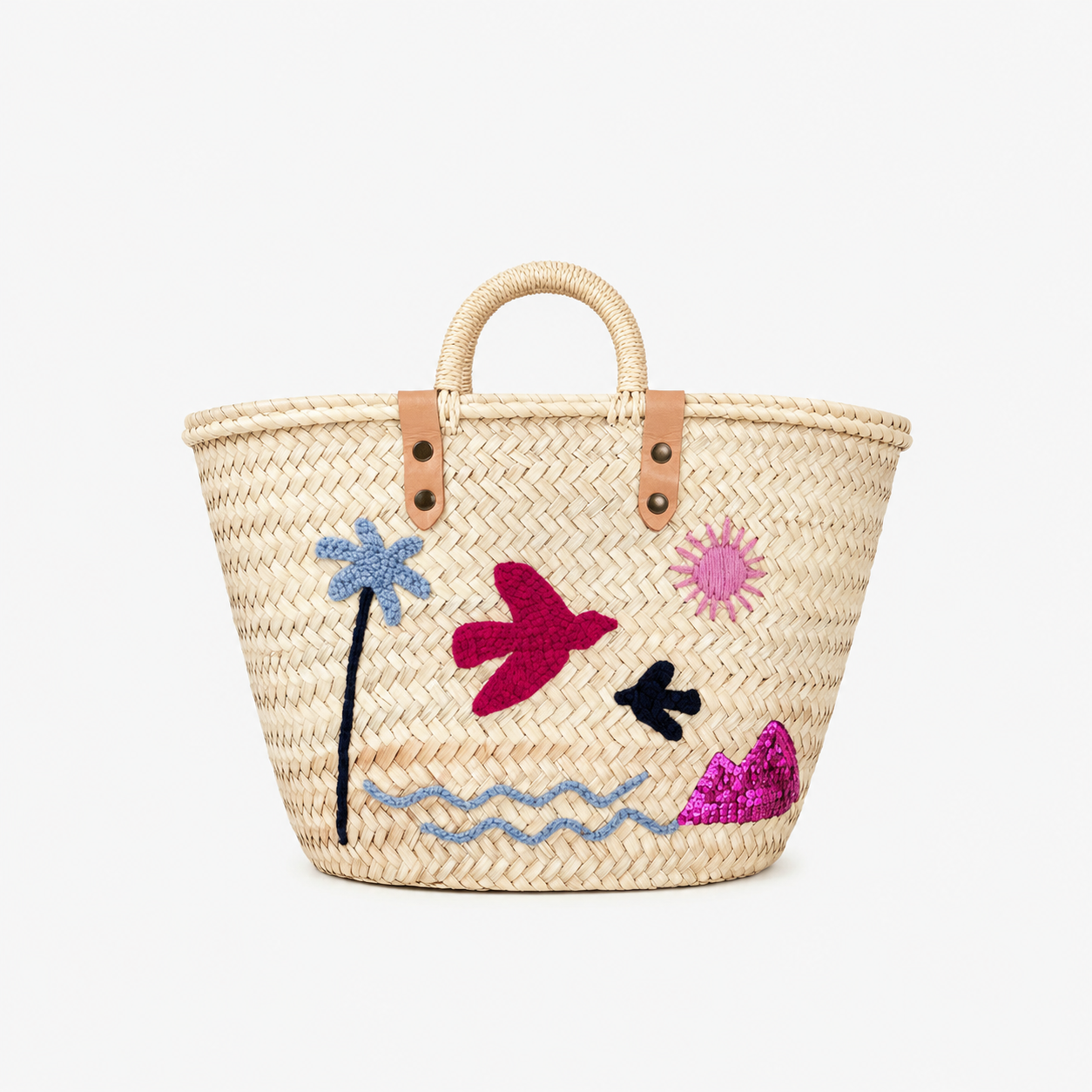 A natural woven straw tote bag with tan leather handles and embroidered tropical motifs including a blue palm tree, pink sun, magenta bird, black bird, and wavy blue line art on a white background.