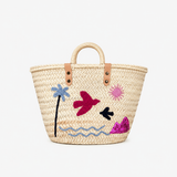 A natural woven straw tote bag with tan leather handles and embroidered tropical motifs including a blue palm tree, pink sun, magenta bird, black bird, and wavy blue line art on a white background.