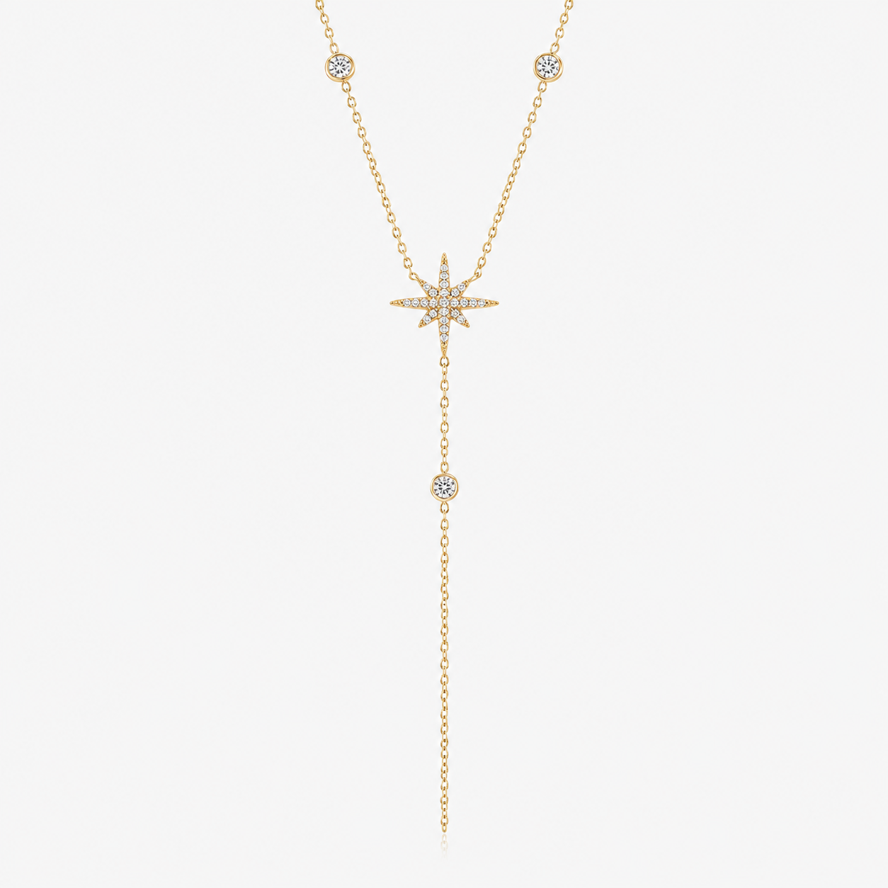 A delicate gold Y-necklace featuring a central starburst cluster of small clear stones, with two drop chains ending in small round stone-set beads, shown on a plain light gray background.