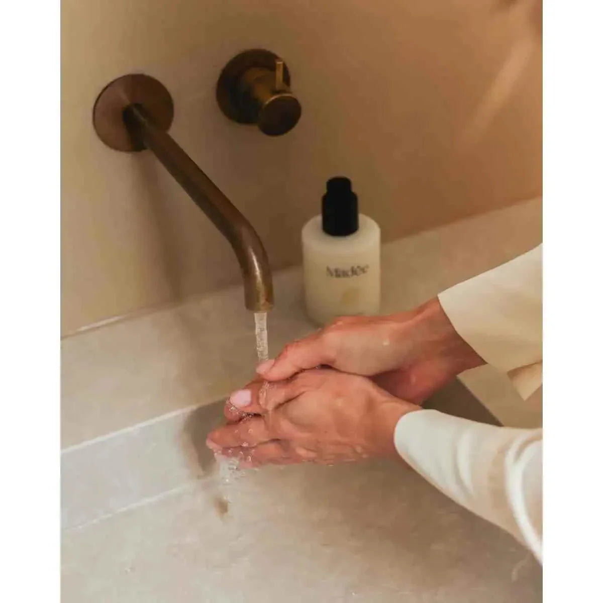 Madée Hand Soap Set with white pump bottle dispensing soap while hands are being washed under a bronze faucet in a beige sink