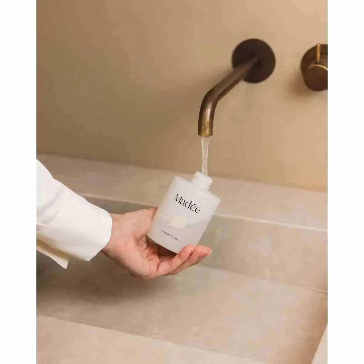Madée Hand Soap Set white minimalist bottle with black text being filled under bronze faucet in beige sink