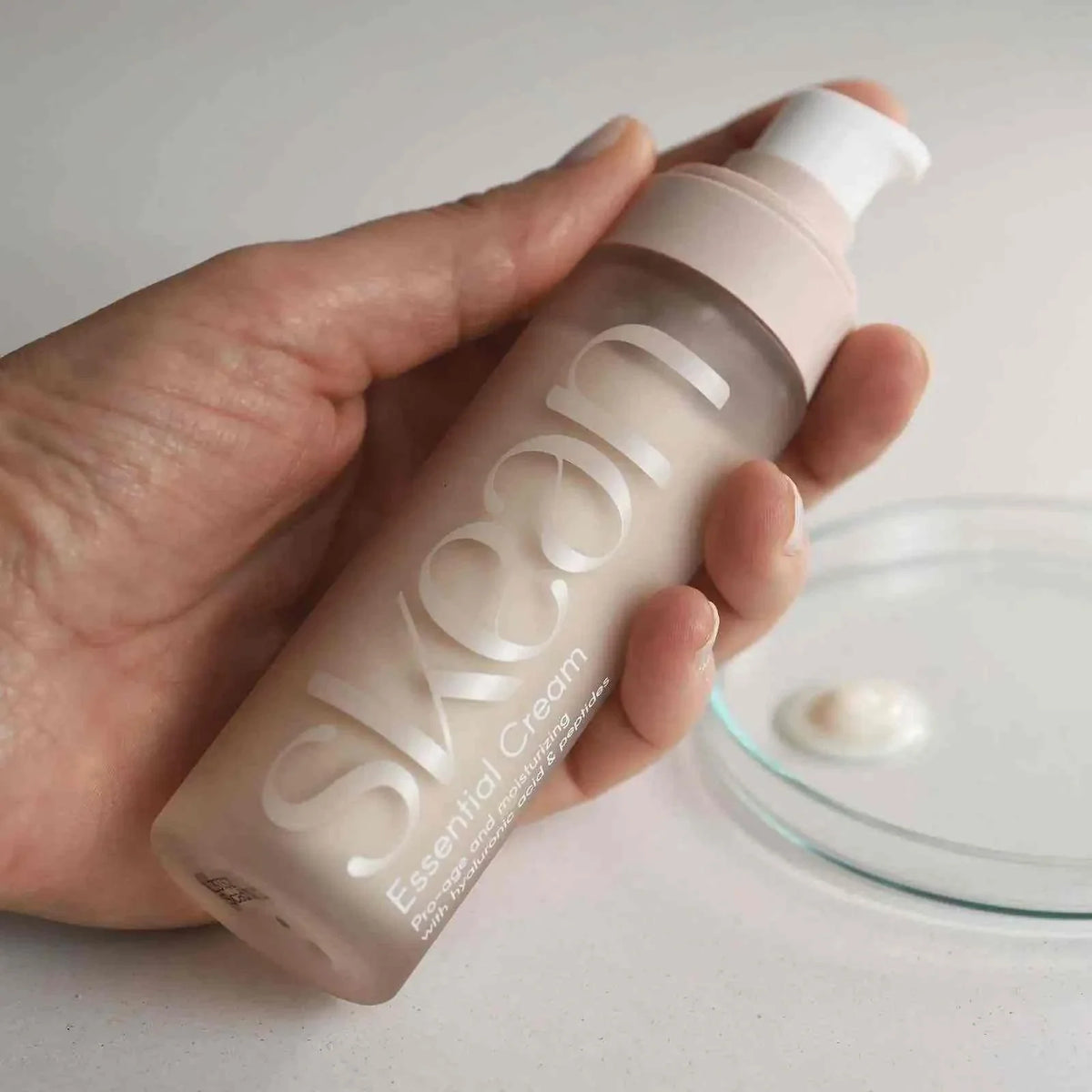 SkeanBeauty Essential Cream in a frosted pink pump bottle held in hand with cream dollop on clear dish
